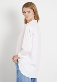 Camisa blanca de botones con mangas largas, con un fit relajado, dobladillo curvado y tejido suave y liso, combinada con jeans de color azul claro.