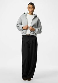 Giovane con capelli ricci che indossa una felpa grigia chiara con zip su una maglietta bianca e pantaloni neri larghi, in piedi davanti a uno sfondo neutro.