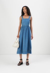 Denim midi dress with square neckline, gathered waist, and wide straps. Black strappy sandals and a small white handbag complete the look.