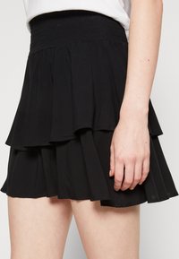Černá mini sukně s volným volánovým páskem, kterou nosí osoba stojící s jednou rukou u boku.