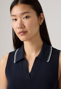 Haut sans manches bleu marine avec un tricot texturé, présentant un col en V et un col à rayures blanches contrastantes. Des bijoux d'oreille sont également visibles.