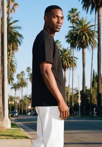 Mann in schwarzem T-Shirt und weißer Hose, der auf einer von Palmen gesäumten Straße unter klarem blauem Himmel steht und über die Schulter schaut.