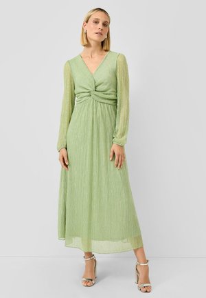 Femme portant une robe midi à manches longues vert clair avec un détail de taille torsadée et des sandales à talons hauts argentées, debout devant un fond uni.