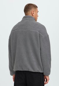 Graue Fleecejacke mit hohem Kragen, strukturiert und lässiger Schnitt. Mit langen Ärmeln und einer Nahtdetail über den Schultern.