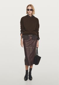 Junge Frau in schwarzer Sonnenbrille, die einen dunkelbraunen Pullover, ein dunkles Shirt, einen ledernen Midirock, schwarze Socken und Schuhe trägt und eine schwarze Tasche hält.