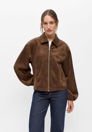 Mujer con una chaqueta de ante marrón con cremallera y mangas largas y vaqueros azules, de pie sobre un fondo blanco liso.
