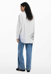 Camicia bianca a maniche lunghe con stampa floreale sul retro, abbinata a jeans larghi blu chiaro e scarpe slip-on nere.