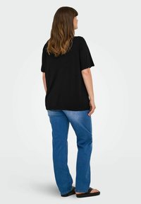 Svart oversized t-shirt med korta ärmar, tillsammans med ljusblå raka jeans och svarta platta sandaler. Modellen står vänd mot vänster.