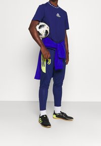 Camiseta atlética de la marina con el logo de Adidas, combinada con pantalones a juego que presentan acentos amarillos. Zapatillas de fútbol negras con detalles neón. Sosteniendo un balón de fútbol.