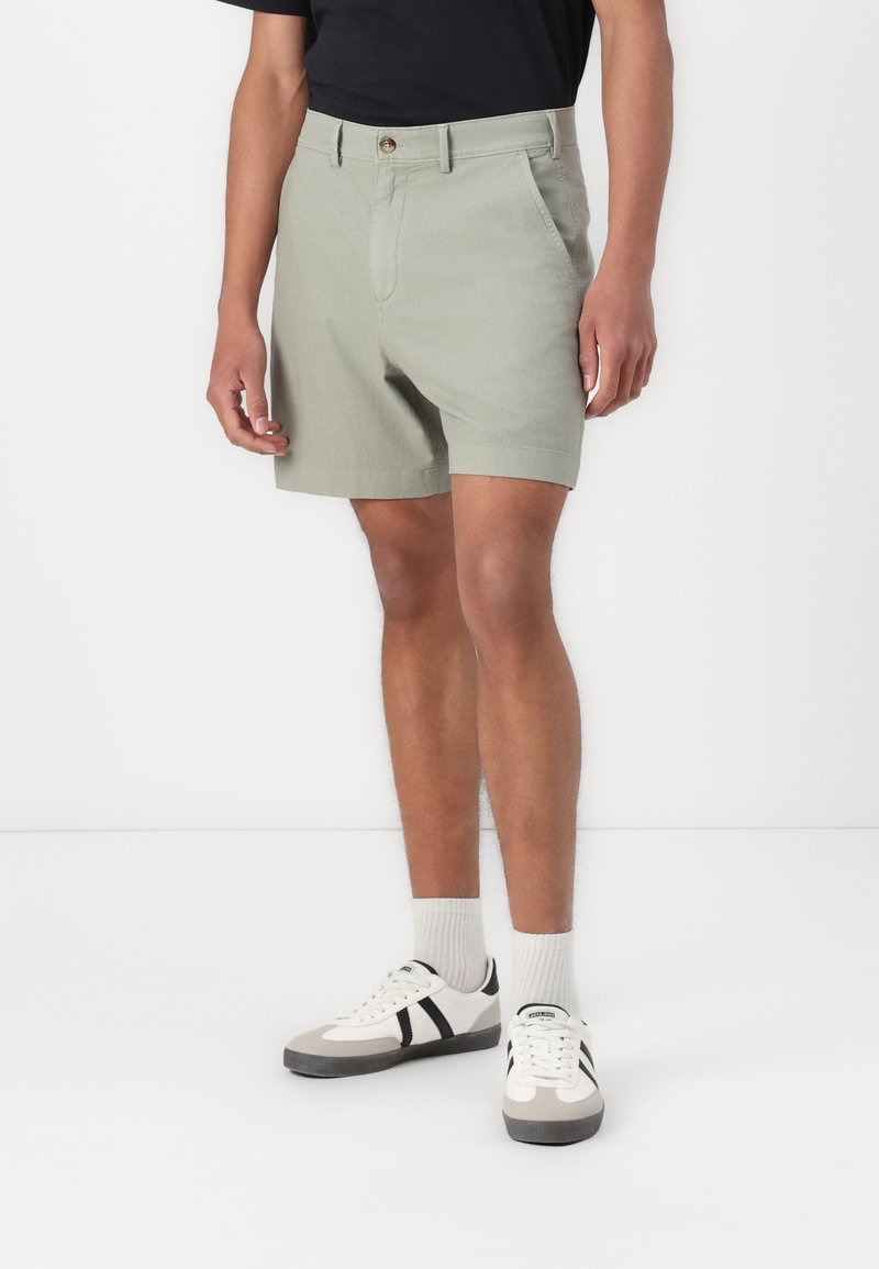 Pantaloni scurți kaki din bumbac, cu front plat, două buzunare laterale și închidere cu un singur nasture. Purtați cu adidași albi și șosete gri.