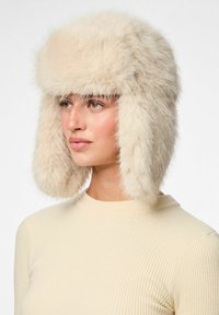 Chapeau en fausse fourrure de couleur beige clair avec des rabats d'oreilles, texture duveteuse et sommet arrondi. Porté par un modèle avec un haut à côtes de couleur crème.
