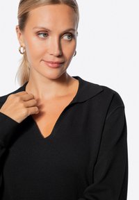 Černý pletený top s výstřihem do V a límečkem. Dlouhé rukávy, ribbovaná struktura a volný střih. Ruka modelky je položena na látce.