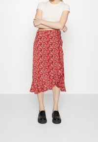 Jupe midi florale rouge en tissu léger et fluide. Présente un ourlet volanté et un lien à la taille pour un look ajusté. Associée à des chaussures noires.
