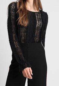 Schwarzes Langarmoberteil mit durchsichtiger Spitze, das strukturierte Streifen und geraffte Bündchen aufweist. Kombiniert mit hochtaillierter schwarzer Hose, die eine figurbetonte Silhouette schafft.