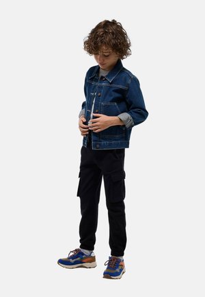Veste en denim avec fermeture à boutons, deux poches avant et poignets gris ; associée à un pantalon cargo noir et des baskets multicolores.