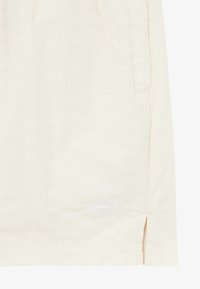 Light yellow shorts made from soft fabric, featuring a subtle all-over logo pattern, side pockets, and a small embroidered logo on the hem.