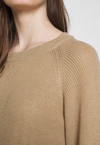 Sweter dziany w ciepłym beżowym kolorze z prążkowanym dekoltem i teksturowanym materiałem. Posiada opuszczone ramiona dla swobodnego kroju.