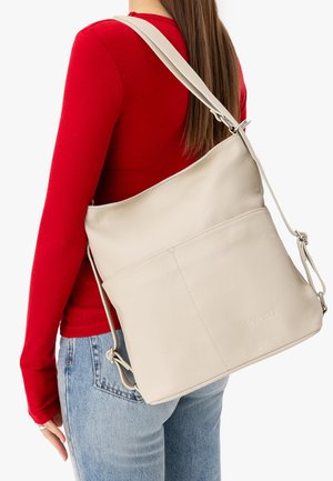 Nő piros hosszú ujjú felsőben és farmerral, nagy bézs színű bőr válltáskát visel a hátán, amely állítható pánttal rendelkezik.