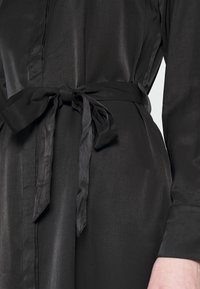 Abito chemisier nero con una texture liscia, caratterizzato da una vita segnato da una cintura e maniche lunghe. Il tessuto ha una leggera lucentezza e un design classico.