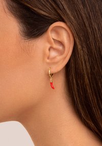 Goldener Creolen-Ohrring mit einem kleinen roten Chilischoten-Anhänger, getragen am rechten Ohr einer Frau vor neutralem Hintergrund.
