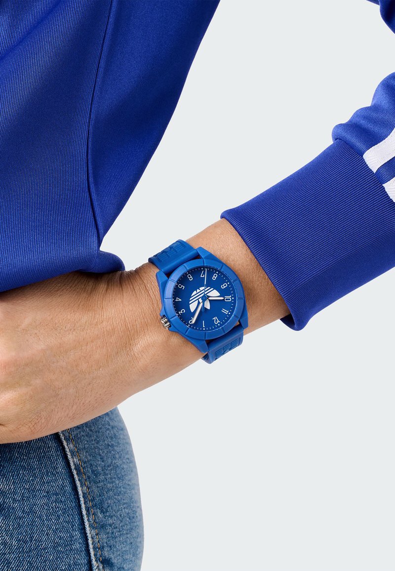 Montre bleue en silicone avec un cadran rond, de grands chiffres blancs et un bracelet texturé, portée sur un poignet à côté d'un vêtement à manches longues bleu.