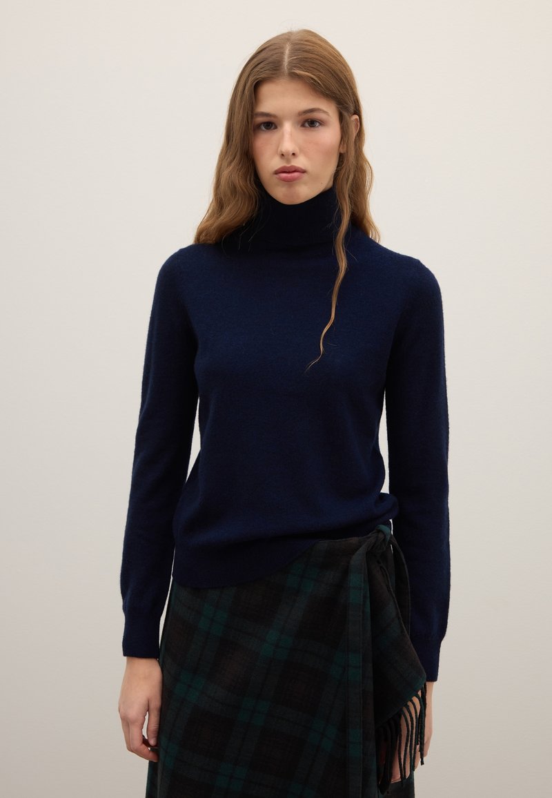 Junge Frau mit langen, welligen Haaren, die einen marineblauen Rollkragenpullover und einen dunkelgrünen und schwarzen karierten Wickelrock trägt.