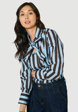 Frau mit langen dunklen Haaren, die eine braun-blau gestreifte Bluse mit Schleifenkragen und dunkelblaue Jeans vor einfarbigem Hintergrund trägt.
