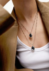 Dos collares de plata con piedras ovaladas negras, uno más largo que el otro, usados sobre una blusa blanca y bajo una chaqueta marrón.