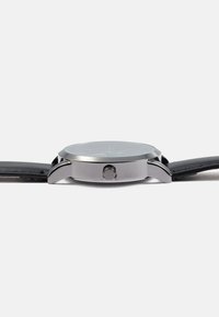 Zegarek ze stali nierdzewnej z okrągłą tarczą, czarnym skórzanym paskiem i boczną koronką. Zegarek charakteryzuje się minimalistycznym designem z wyraźnymi oznaczeniami.