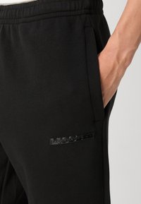Pantalons de survêtement noirs en tissu texturé, avec une poche latérale et une patch logo "LACOSTE" noir brillant sur la cuisse.