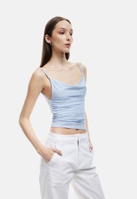 Vaaleansininen, vartaloa myötäilevä hihaton toppi ohuilla olkaimilla, jossa on hienovarainen kukkakuosi ja kerrostettu pinta, yhdistettynä valkoisiin leveälahkeisiin housuihin.