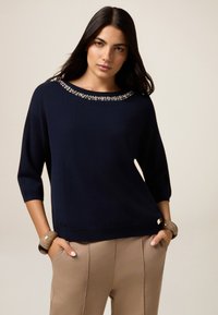 Pull bleu marine avec des manches trois-quarts et un col orné de perles. Le tissu est doux avec une texture lisse. Associé à un pantalon beige clair.
