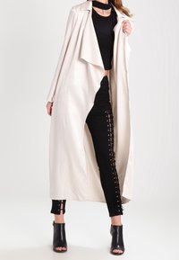 Dlouhý béžový cardigan vyrobený z lehkého materiálu, vrstvený přes černý crop top a černé kalhoty s šněrovacím detailem po stranách. Černé lodičky s otevřenou špičkou.