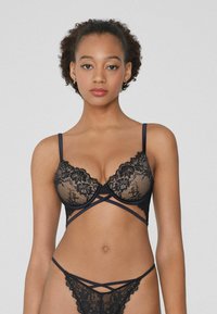 Conjunto de lencería de encaje negro que incluye un sujetador con realce y tirantes en forma de cruz, además de un tanga a juego. Patrón de encaje detallado y acabado suave.