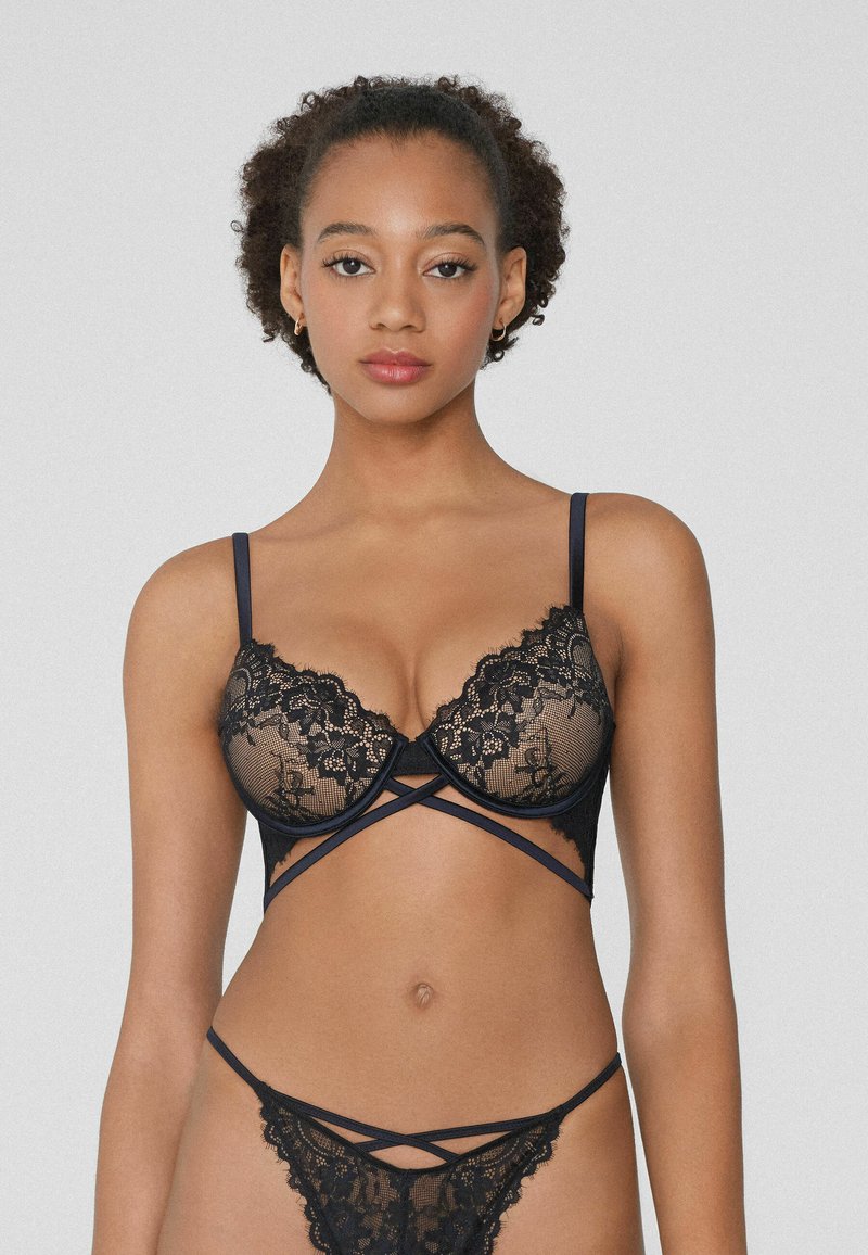 Conjunto de lencería de encaje negro que incluye un sujetador con realce y tirantes en forma de cruz, además de un tanga a juego. Patrón de encaje detallado y acabado suave.