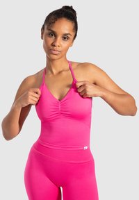 Donna che sistema le cinghie di un top atletico senza maniche di un brillante rosa, indossando leggings a vita alta abbinati, in piedi contro uno sfondo semplice.