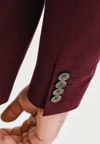 Rękaw burgundowej marynarki z teksturowanej tkaniny; posiada cztery okrągłe guziki z wygrawerowanym logo, podkreślające dopasowany krój.