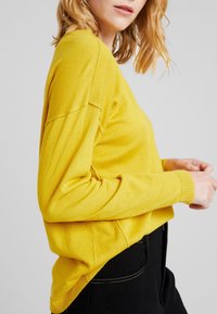 Geel gebreide trui met een relaxte pasvorm, afgeplatte schouders en geribbelde manchetten. Gepaard met zwarte broek met zichtbare stiksels.