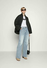Chaqueta negra acolchada y oversize, camiseta gráfica blanca, vaqueros de pierna ancha azul claro, botas beige hasta el tobillo, bolso negro y gafas de sol oscuras.