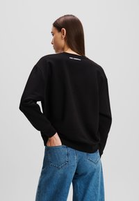 Schwarzer Pullover mit Rundhalsausschnitt und gerippten Bündchen, mit dezentem weißen Logo auf der Rückseite. Kombiniert mit hellblauen Jeans.