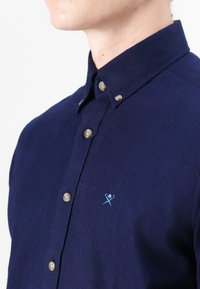 Camisa de botones azul marino hecha de tela texturizada, con logo bordado en azul claro y botones beige en la tapeta.