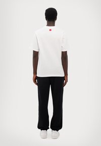 Hvid bomulds t-shirt med et rødt grafik på ryggen, parret med sorte bukser med snøre og hvide sneakers, set fra ryggen.