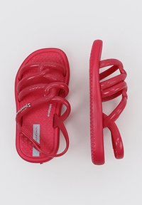 Sandalias de plástico rojo con tres correas en bucle y una plantilla texturizada. Presentan bordes suaves y una suela flexible. Incluyen una correa ajustable para el tobillo.