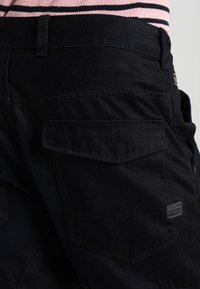 Pantalones cortos de mezclilla negra con una cintura ajustada, múltiples bolsillos y un pequeño parche con logo en la parte trasera. Presentan una textura suave y costuras robustas.