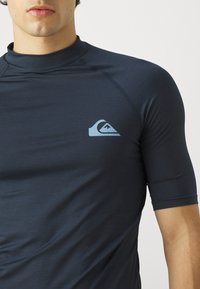 Granatowa koszulka rash guard z krótkim rękawem, wykonana z teksturowanej tkaniny, z jasnoniebieskim logo na lewym piersi. Wysoki kołnierz.