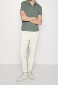 Polo en vert olive avec un col zippé, associé à un pantalon slim-fit blanc cassé et des baskets gris clair. Design simple et moderne.