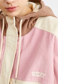 Farverig kordfløjljakke med et pink, creme og brunt design. Har en hætte, snore og et broderet "ROXY" logo på kanten.
