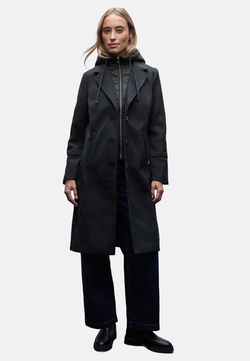 Zwart wollen blend jas met een getailleerde snit, dubbelgebonden ontwerp, capuchon, zijzakken en gestructureerde afwerking, gecombineerd met een donkere spijkerbroek.