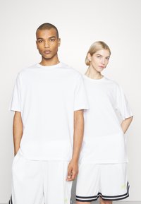 Fehér pamut t-shirt-ek rövid ujjakkal, laza fazonnal és kerek nyakkivágással, fehér nadrággal viselve, melynek fekete és neon zöld részletei vannak.