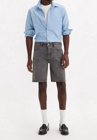 Hombre que lleva una camisa de botón azul claro, camiseta blanca, pantalones cortos de mezclilla grises, calcetines blancos y mocasines de cuero negros frente a un fondo neutro.